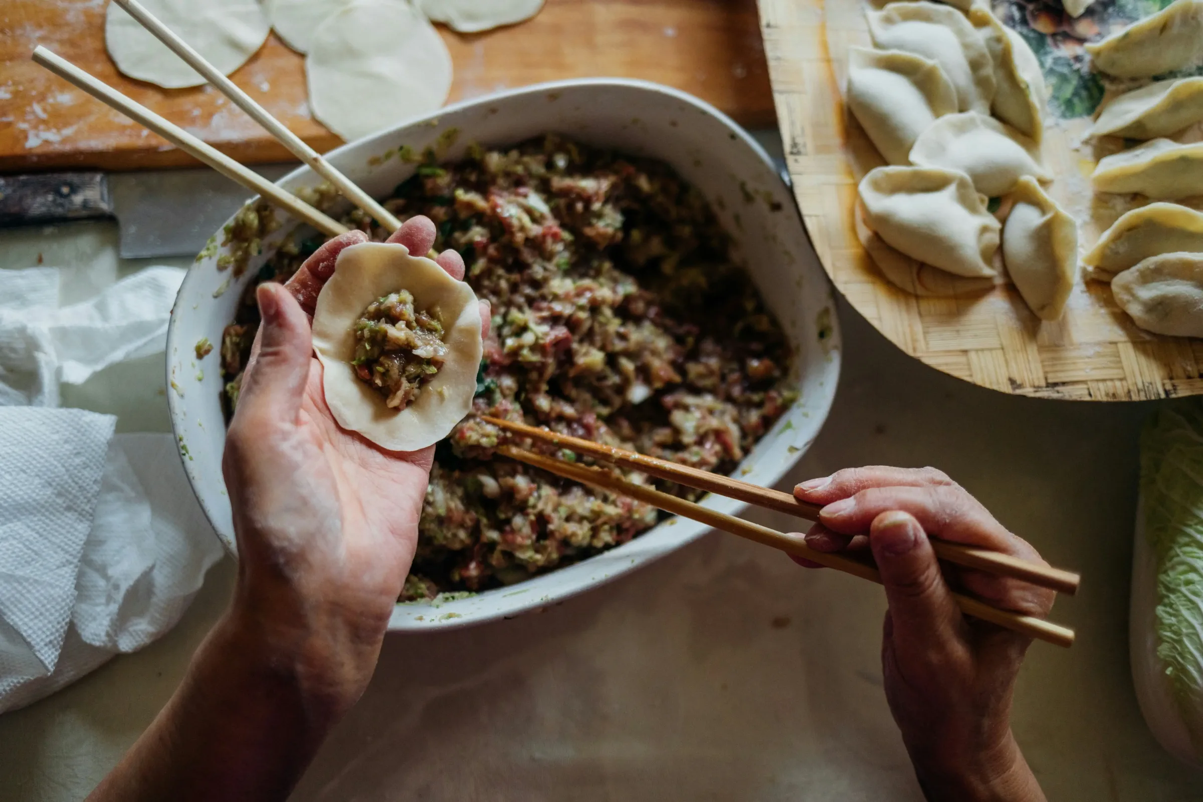 Hands folding a wonton over a bowl of pork and cabbage filling, with a tray of finished dumplings beside it